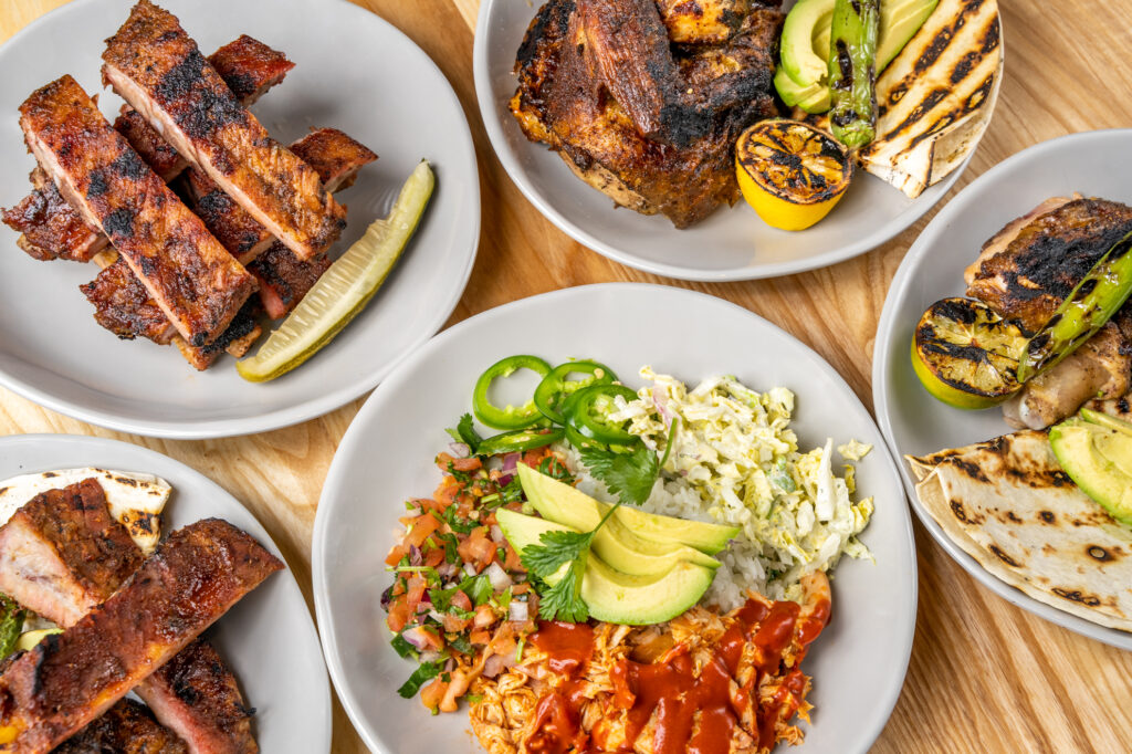 Five plates of food on a wooden table, featuring grilled ribs, roast chicken, grilled vegetables, rice with avocado and salsa, coleslaw, and quesadillas, garnished with jalapeño slices and fresh herbs.