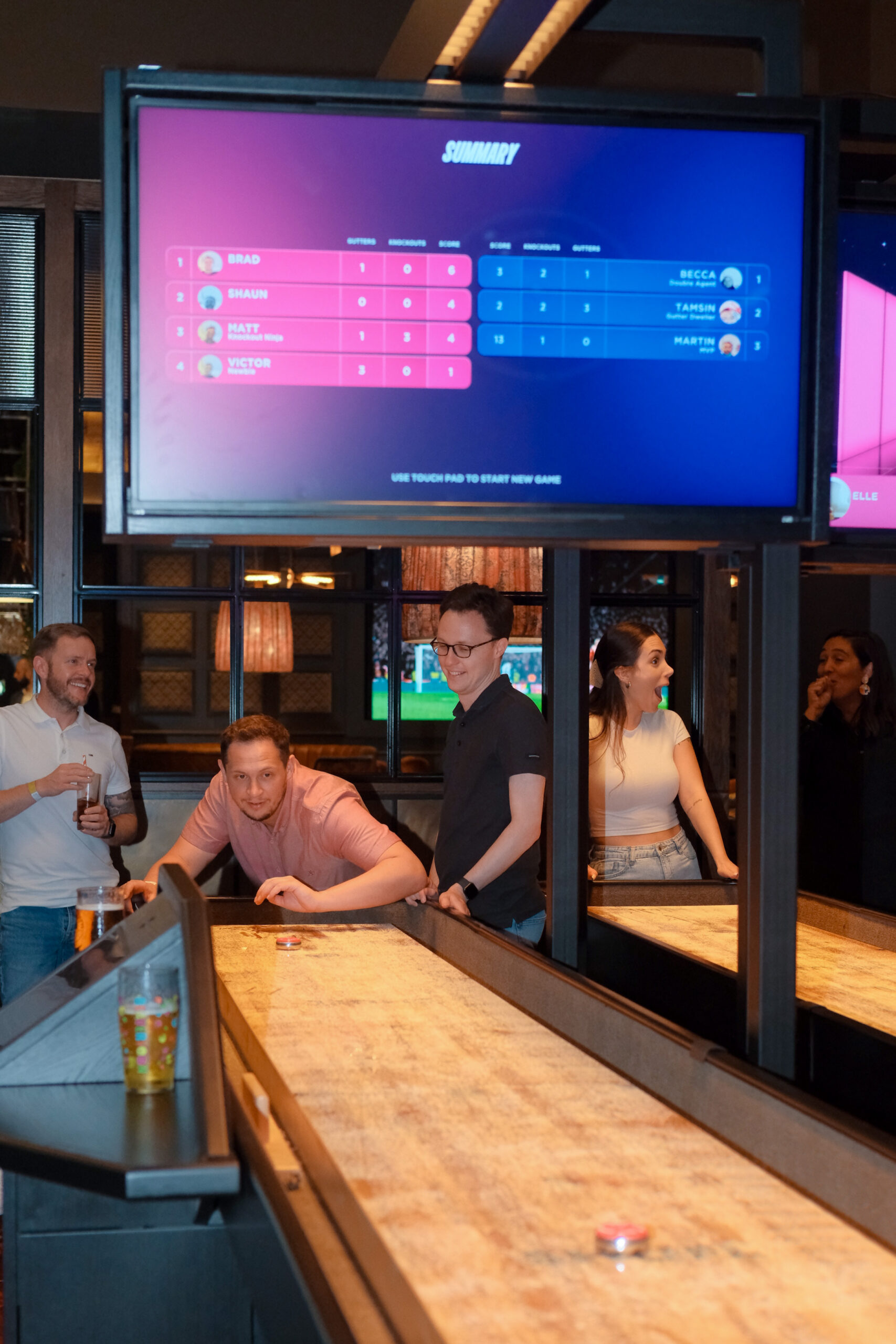 Four people play shuffleboard indoors. Two are focused on the game, while the other two watch and smile. A scoreboard on a screen above shows players names and scores. Drinks sit on the edge of the shuffleboard table.