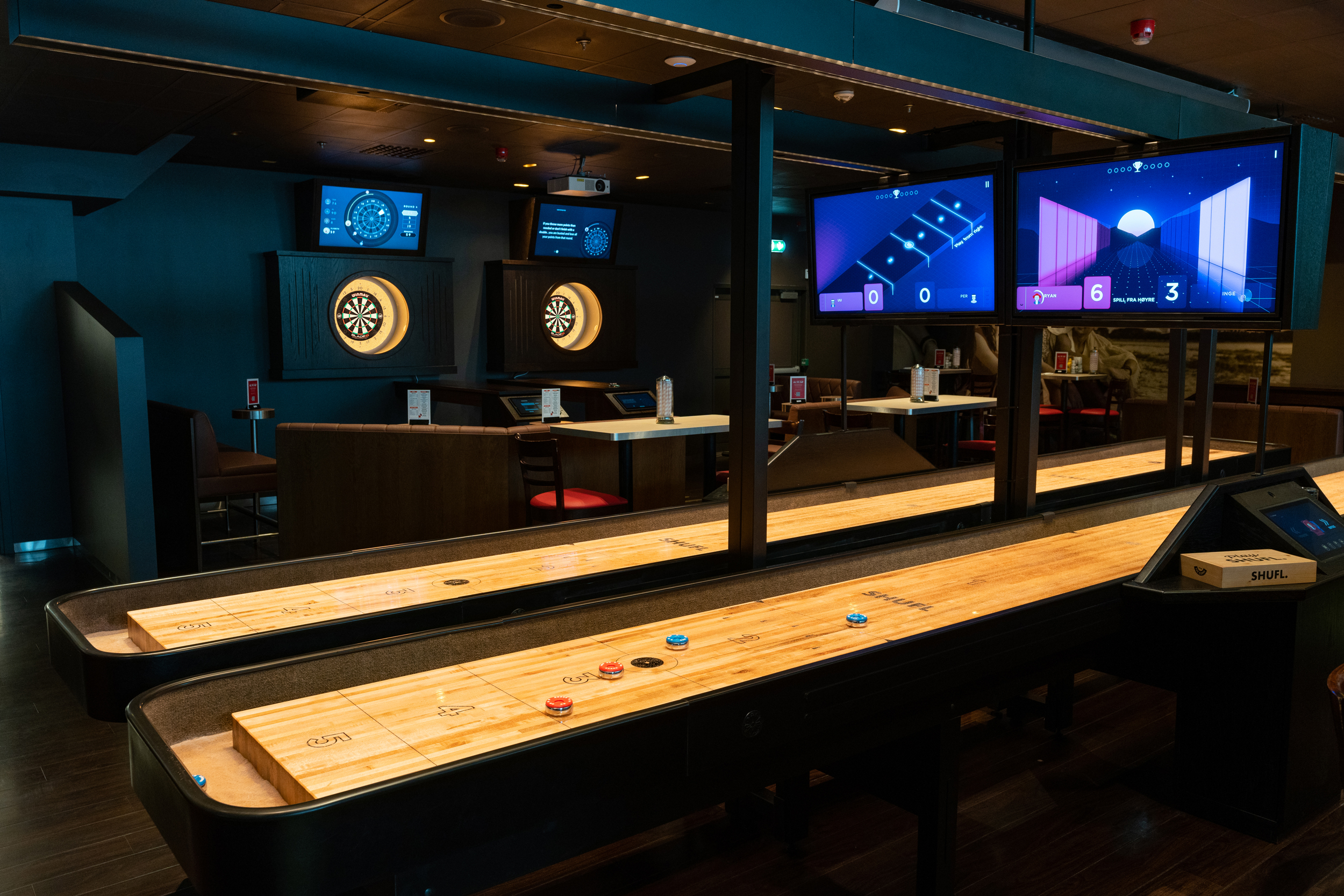 A game room with two shuffleboard tables in the foreground and electronic dartboards with score screens in the background. The lighting is dim, creating a modern, relaxed atmosphere.