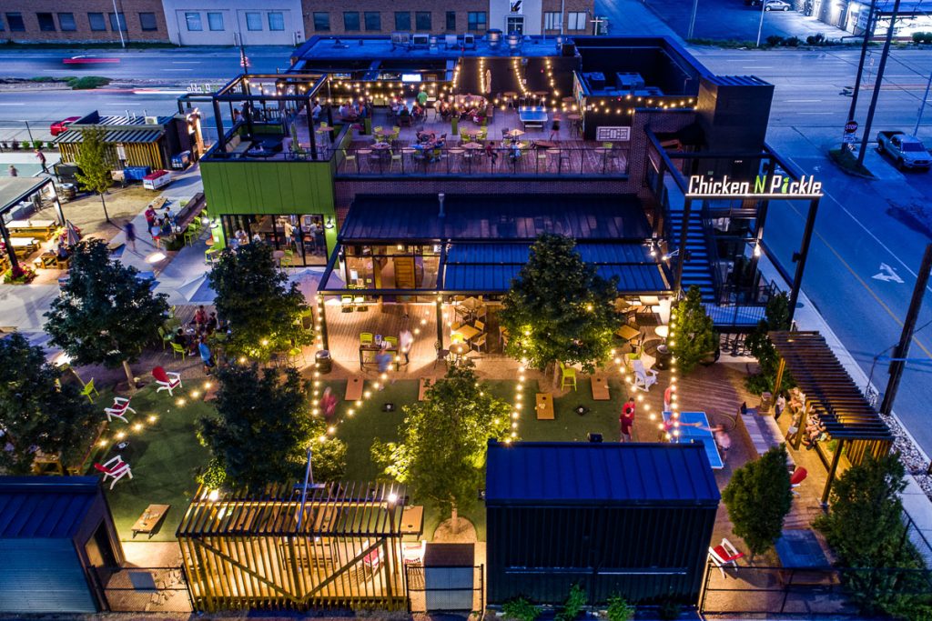 Aerial view of Chicken N Pickle at dusk, with string lights illuminating outdoor seating, pickleball courts, and picnic tables surrounded by trees and greenery, creating a lively, festive atmosphere.