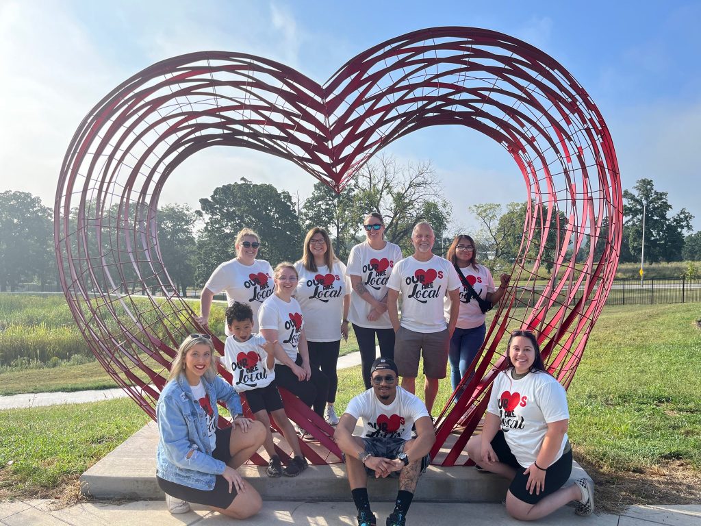 Nine people wearing matching Love Local shirts pose and smile in front of a large red heart-shaped metal sculpture in an outdoor park setting on a sunny day.