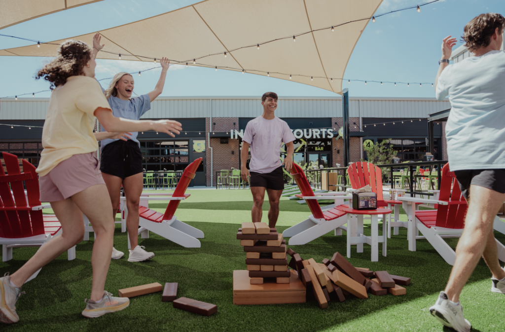 Four young people laugh and react as a giant Jenga tower collapses on a lawn under a canopy, surrounded by red chairs at an outdoor venue.