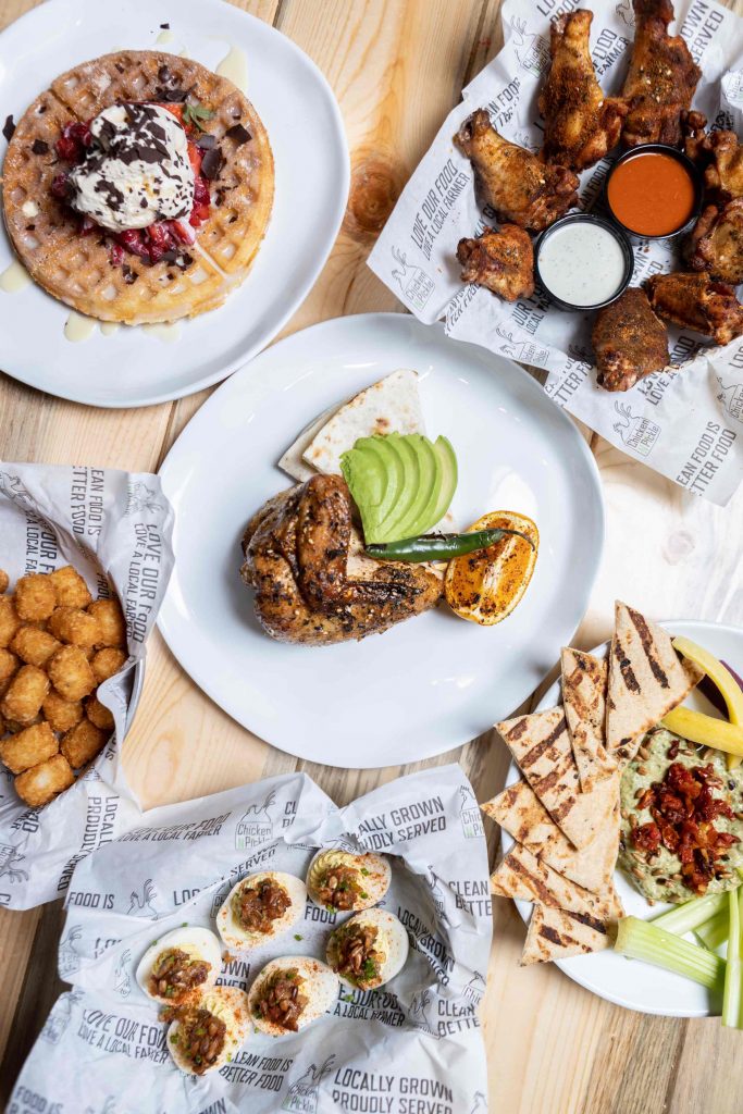 A table with plates of food, including waffles with whipped cream and strawberries, chicken wings with dipping sauces, grilled chicken with avocado, tater tots, pita with hummus, and deviled eggs on parchment paper.
