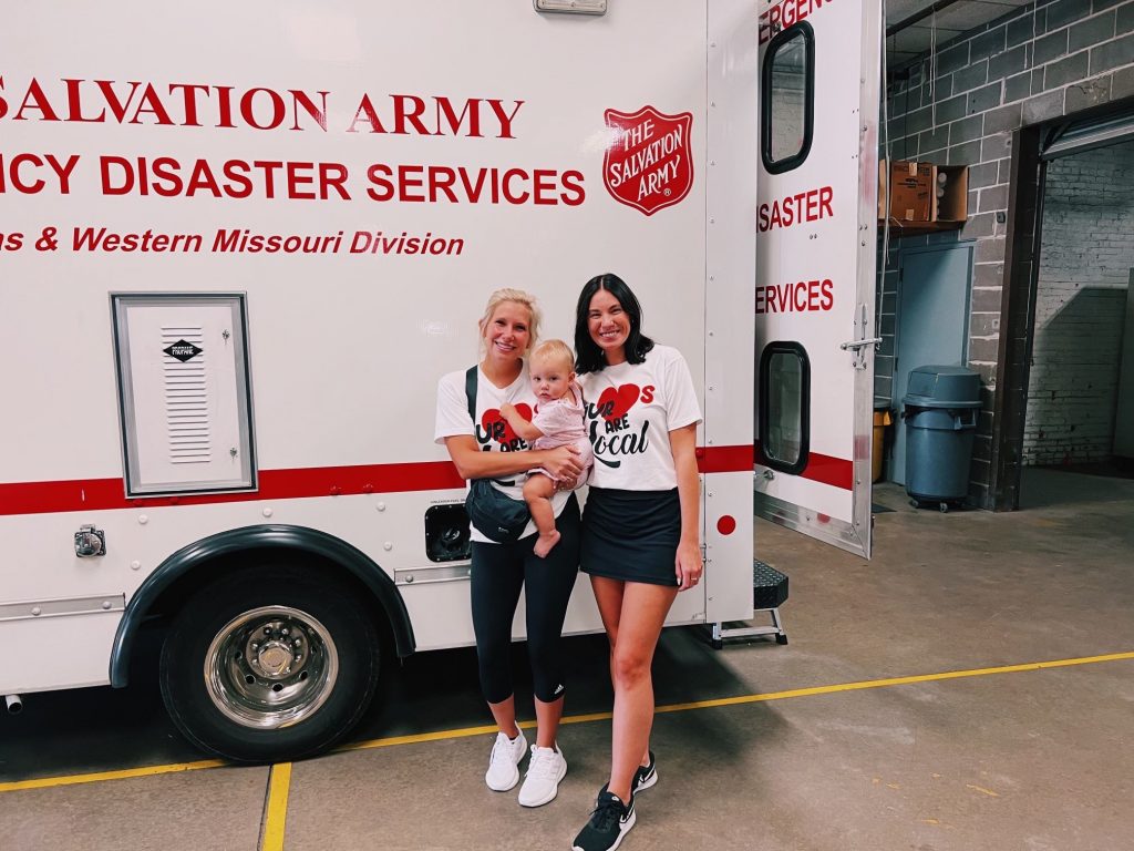 Two women and a baby stand smiling in front of a white Salvation Army Emergency Disaster Services vehicle inside a garage. The women wear matching white T-shirts and black skirts.