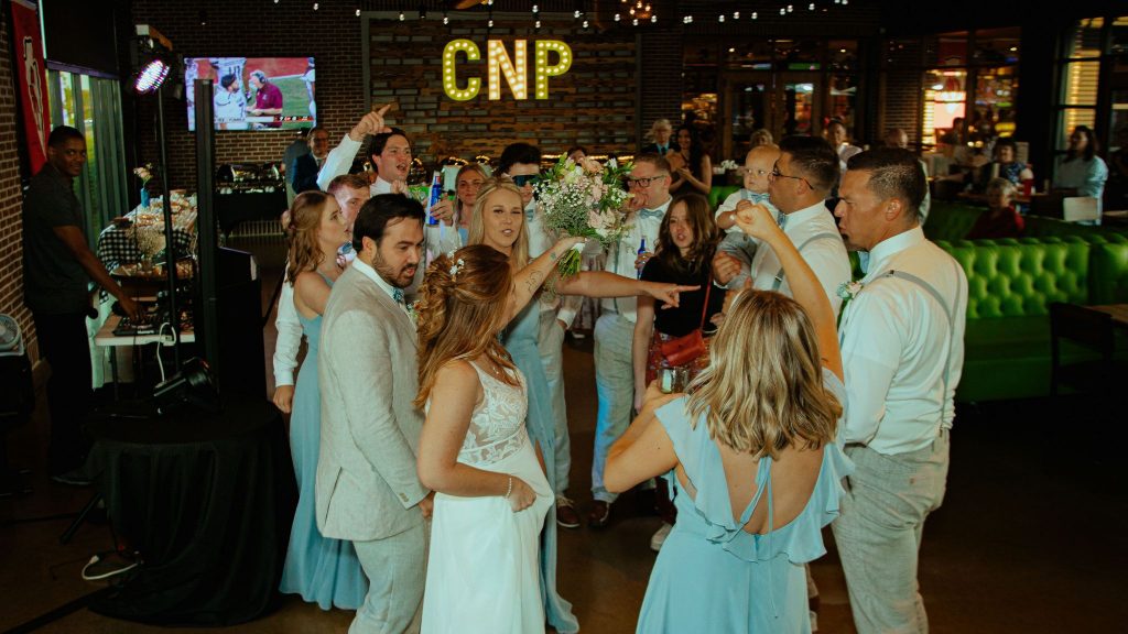 A group of people dressed in formal attire joyfully dance in a circle at an indoor event, with a lit CNP sign and green booth seating visible in the background.