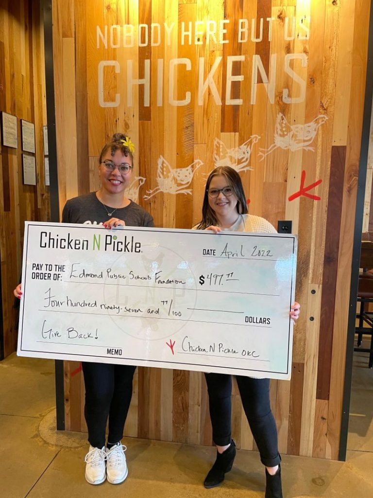 Two women smiling and holding a large donation check for $497.77, made out to Edmond Public Schools Foundation from Chicken N Pickle. Behind them is a wood-paneled wall with the words NOBODY HERE BUT US CHICKENS.