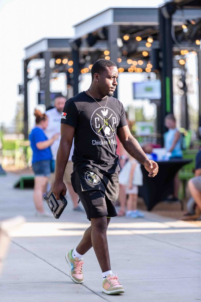 A man in a black Chicken N Pickle t-shirt and shorts walks outdoors, holding a paddle and phone. People and string lights are visible in the blurred background.