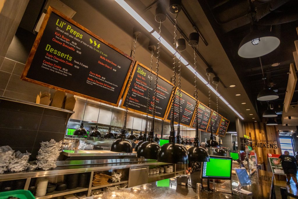 A restaurant counter with hanging menu boards displaying food options, including Lil’ Peeps, desserts, and sides. Pendant lights hang above the counter, with kitchen equipment and green-lit monitors visible below.