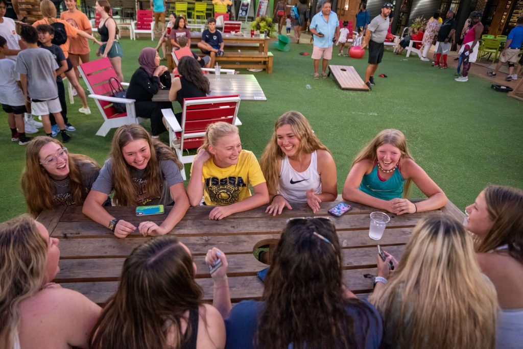 A group of smiling teenage girls sit around an outdoor wooden table, talking and laughing, while other people socialize and children play in a lively, well-lit recreational area with green artificial turf.