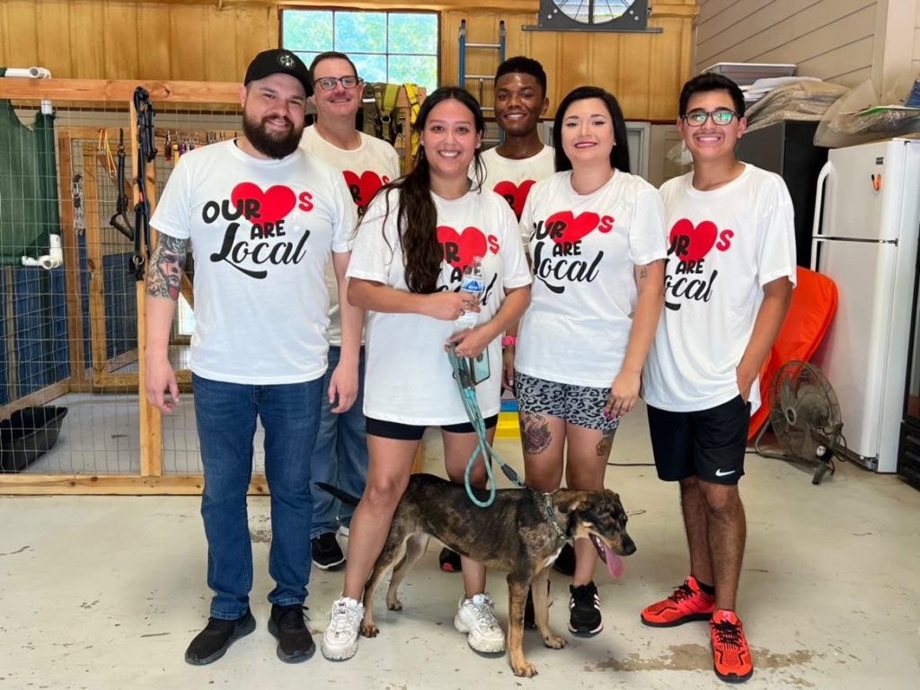 Six smiling people wearing matching Our Hearts Are Local shirts stand together indoors. One woman in the center holds the leash of a happy dog. Shelving, cages, and a fan are visible in the background.