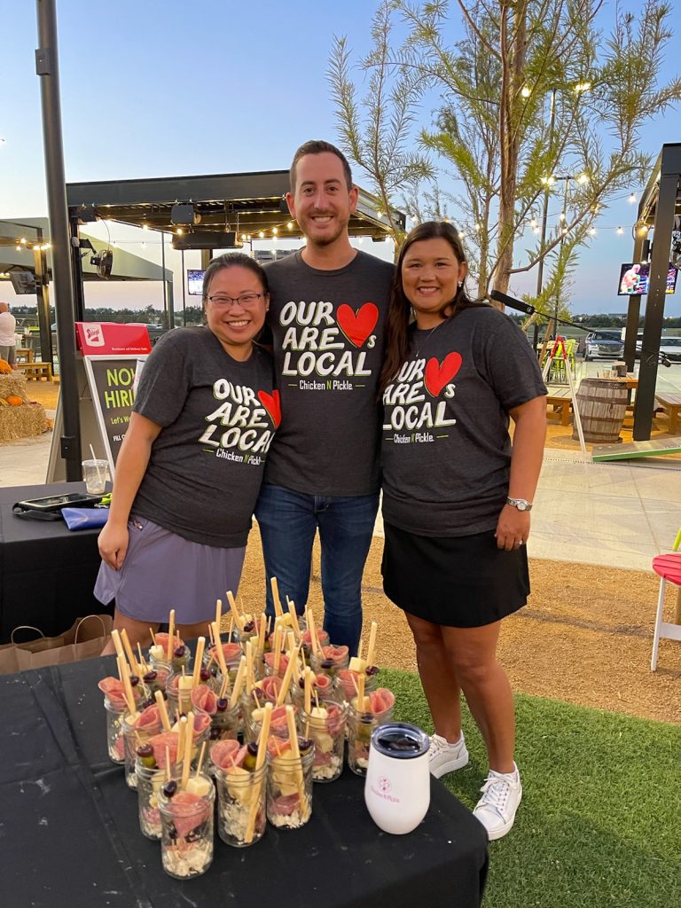 Three smiling people stand behind a table of snack cups at an outdoor event. They wear matching “Our Heart Are Local” t-shirts and pose together in front of trees and vendor booths at sunset.