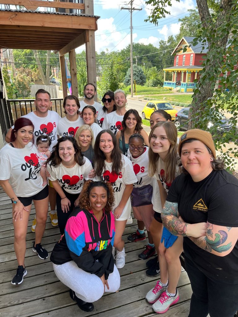 A diverse group of people, some wearing matching Our Roots Local shirts, smile together on a wooden deck outdoors with trees and houses in the background.