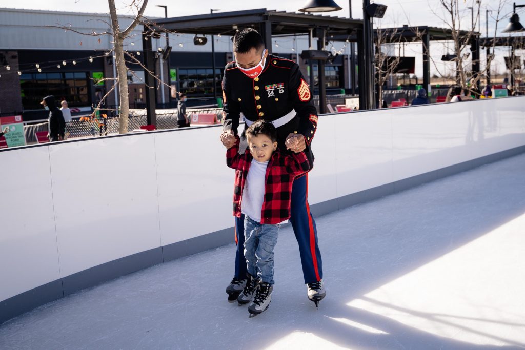 A U.S. Marine in dress uniform helps a young boy in a red plaid jacket ice skate outdoors during the day. They are smiling and holding hands on the rink.