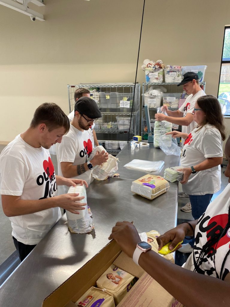 A group of people wearing matching Our Love T-shirts pack diapers into plastic bags around a metal table in a well-lit room with shelves and supplies in the background.