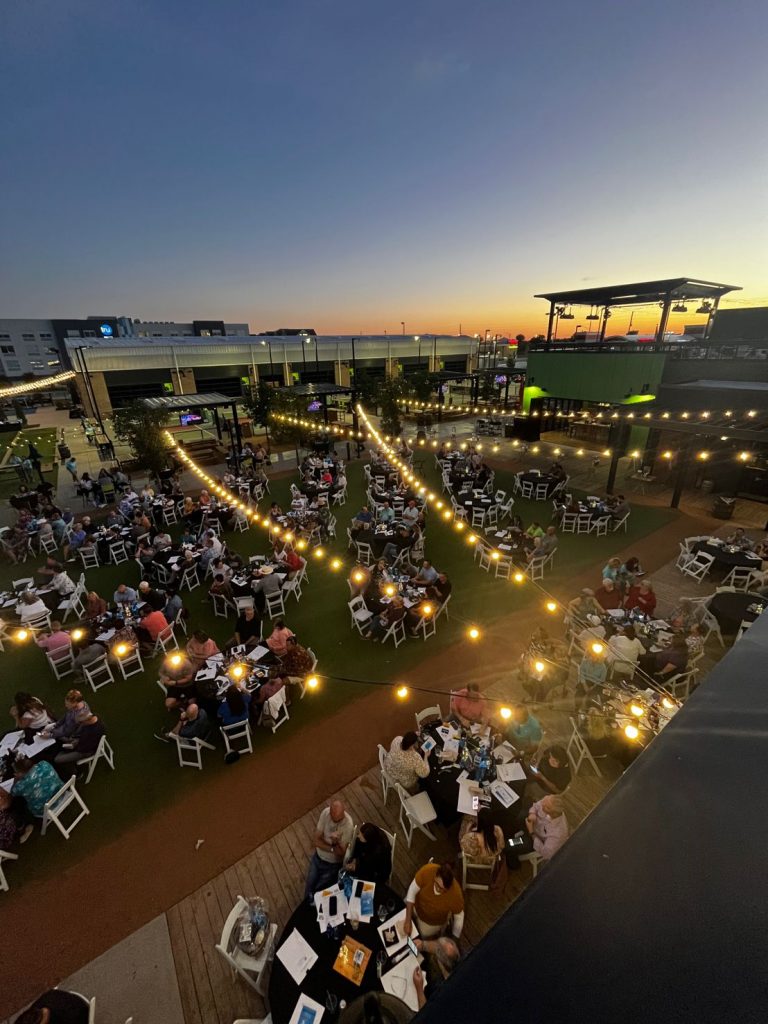 A rooftop outdoor event at sunset with many round tables surrounded by people. String lights are hung above, creating a warm atmosphere. Buildings and an orange sky are visible in the background.