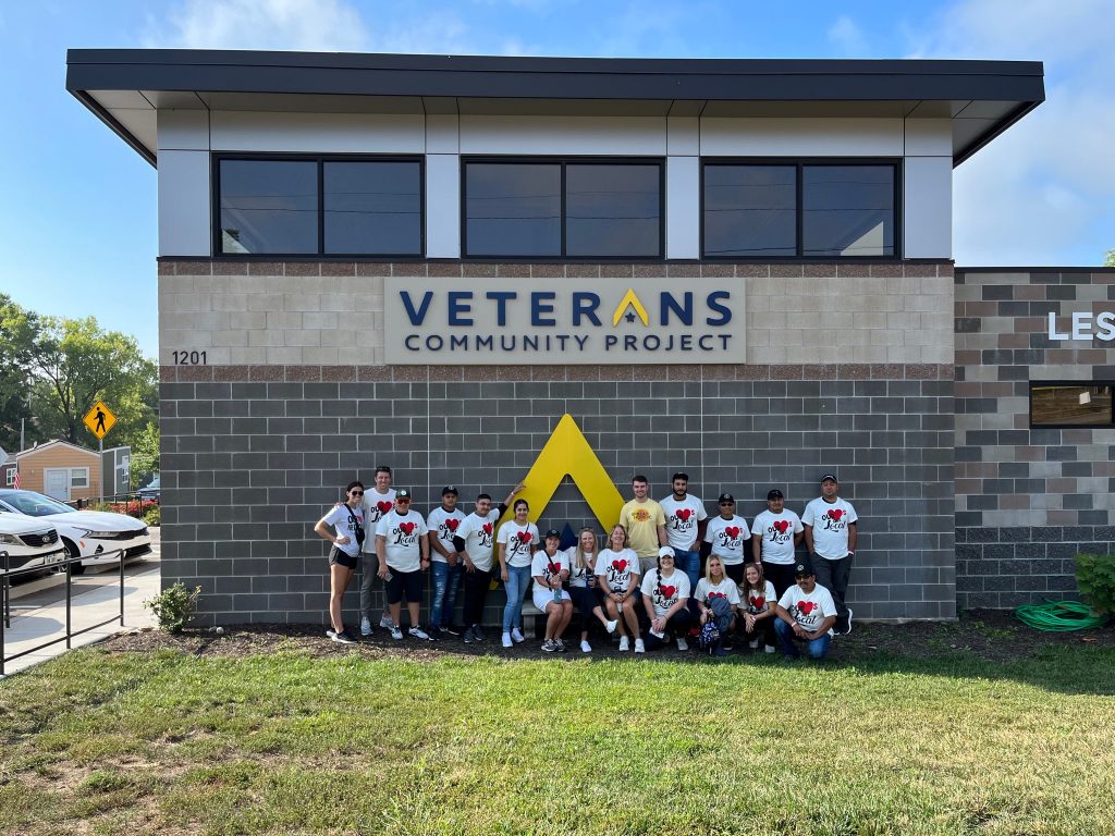A group of people wearing matching white shirts with red designs pose together in front of a building labeled “Veterans Community Project” on a sunny day.