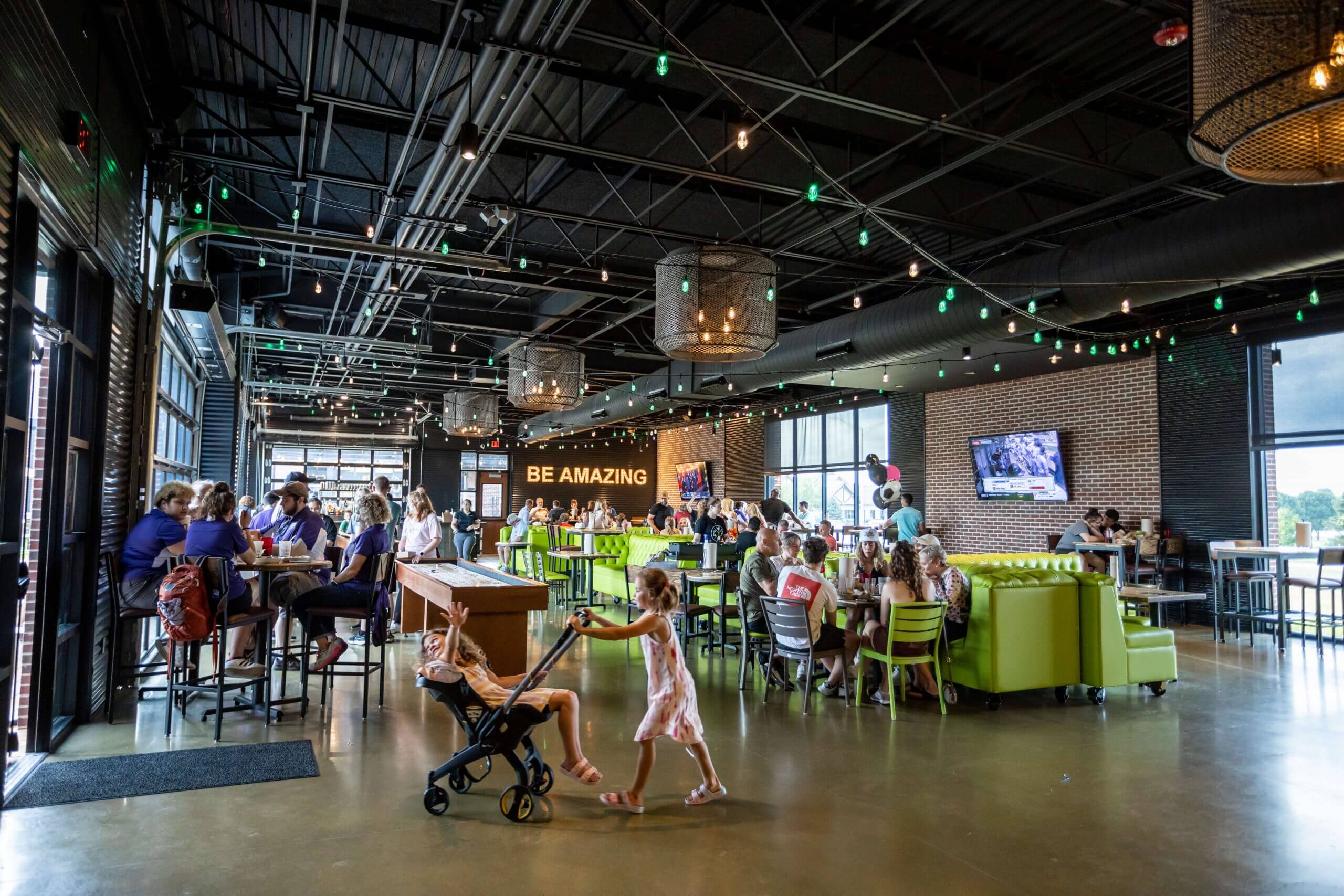 A lively, modern restaurant with green booths and string lights, filled with people dining. A child pushes another child in a stroller across the floor. A neon sign on the brick wall reads BE AMAZING.