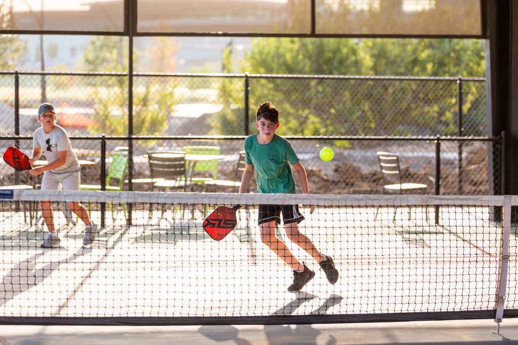Two boys play pickleball on an outdoor court. One boy in a turquoise shirt moves toward the net to hit the ball, while the other stands ready in the background. Sunlight filters through trees and chairs outside the fence.