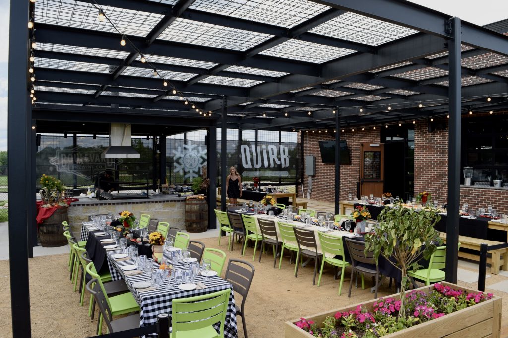Outdoor dining area with two long tables set for a meal under a metal pergola, string lights overhead, potted plants, and a grill station in the background. A brick building and a QUIRK sign are visible.