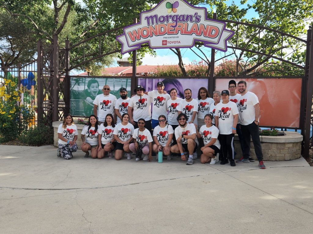 A group of people wearing matching Keep it Real T-shirts pose and smile together in front of the entrance sign for Morgan’s Wonderland, an inclusive amusement park. Trees and colorful banners are in the background.