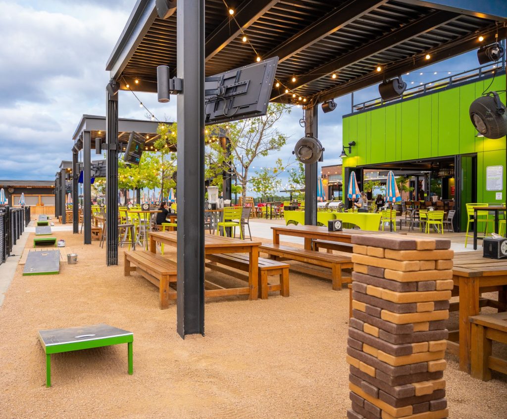 Outdoor dining area with wooden picnic tables, string lights, green chairs, cornhole boards, and a giant Jenga game on gravel, surrounded by modern structures and greenery under a cloudy sky.