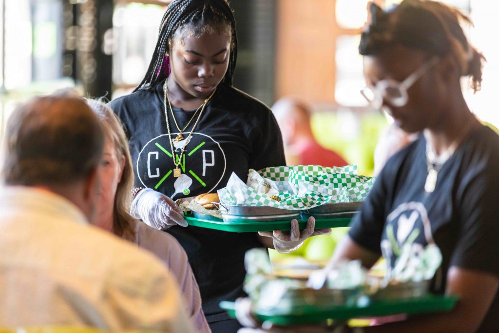 Two servers in black shirts with a heart design deliver trays of food from the North Kansas City Menu to diners. The trays, lined with green and white checkered paper, are topped with delicious sandwiches.