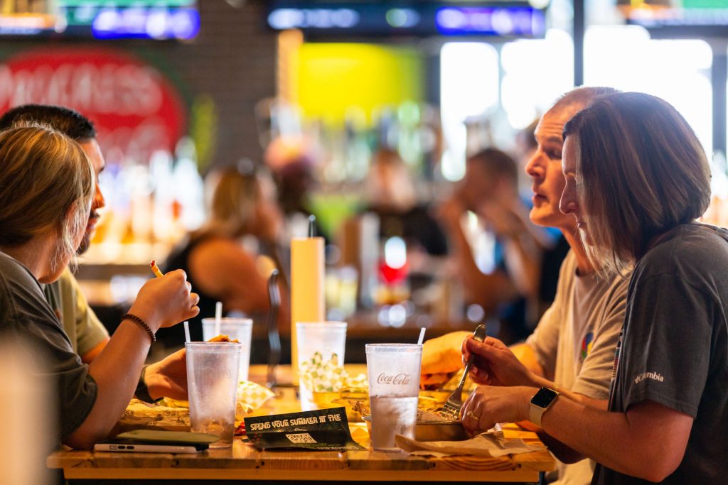 Four people sit at a restaurant table, eating and talking. Glasses of drinks and plates of food are in front of them. The background is blurred, showing a lively, colorful dining area.