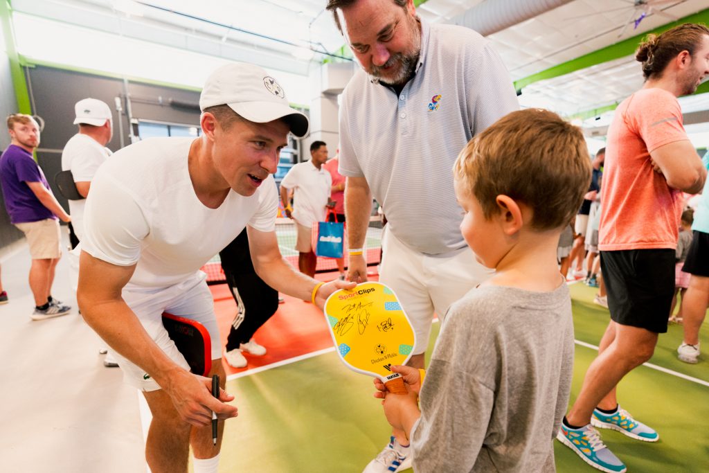 A man in sportswear signs a yellow pickleball paddle for a young boy while others watch in an indoor court. The boy looks at the paddle, and the atmosphere appears lively and friendly.