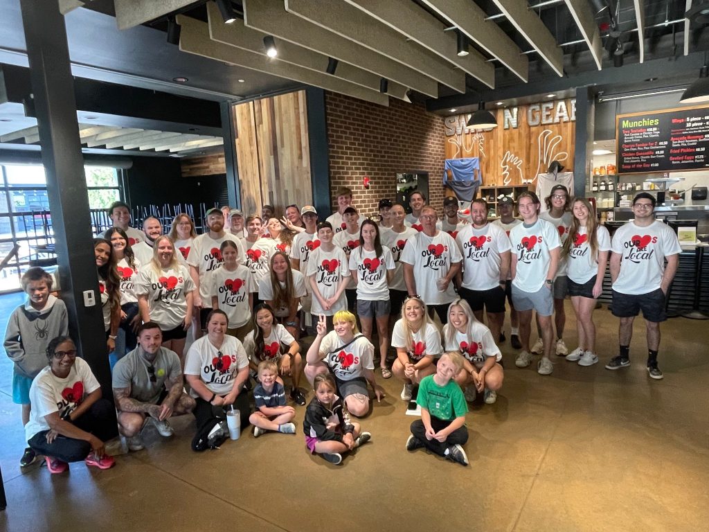A large group of people, many wearing matching white “I Love Leah” shirts, pose and smile inside a casual restaurant with brick walls and menu boards in the background. Some children sit in the front.
