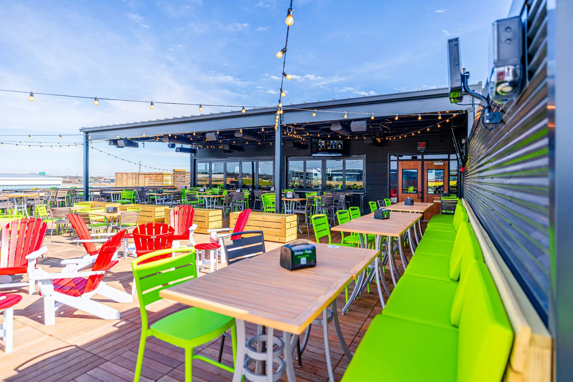 A vibrant rooftop patio with green chairs, wooden tables, red and white Adirondack chairs, string lights, and large windows under a clear blue sky.
