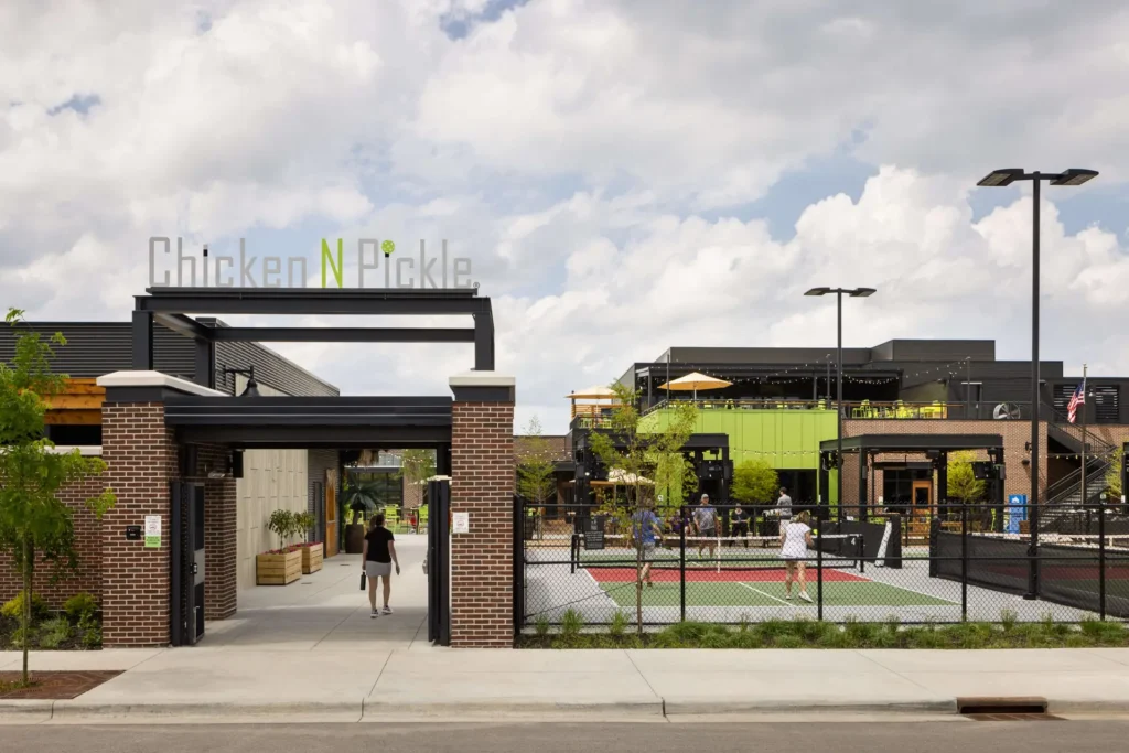Entrance to Chicken N Pickle, an outdoor recreation and dining venue, with people playing pickleball on courts and others walking near modern buildings under a partly cloudy sky.