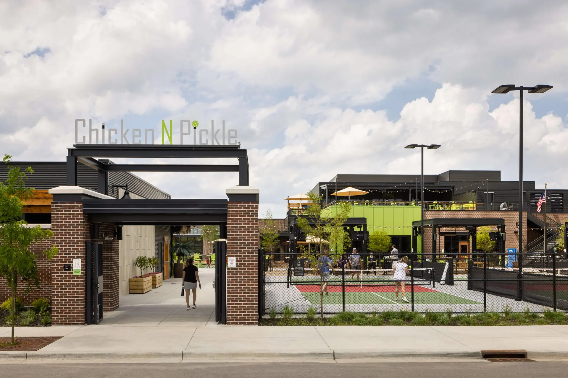 Entrance to Chicken N Pickle, an outdoor recreation and dining venue, with people playing pickleball on courts and others walking near modern buildings under a partly cloudy sky.