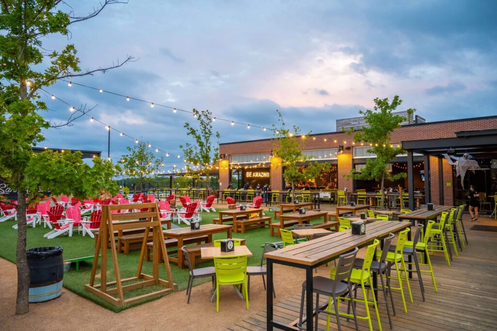 Outdoor patio with string lights, wooden tables, green and yellow chairs, red lounge chairs, and swings on artificial grass. People are seated in the background near a brick building at dusk under a cloudy sky.
