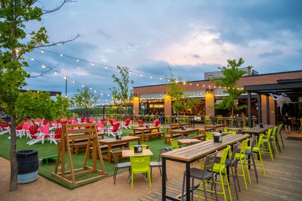 Outdoor patio with string lights, wooden tables, green and yellow chairs, red lounge chairs, and swings on artificial grass. People are seated in the background near a brick building at dusk under a cloudy sky.
