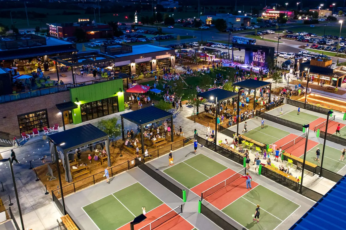 A lively outdoor venue at night with people playing on multiple pickleball courts, others dining on patios, and groups gathered near a stage and grassy area, surrounded by restaurants and parked cars.