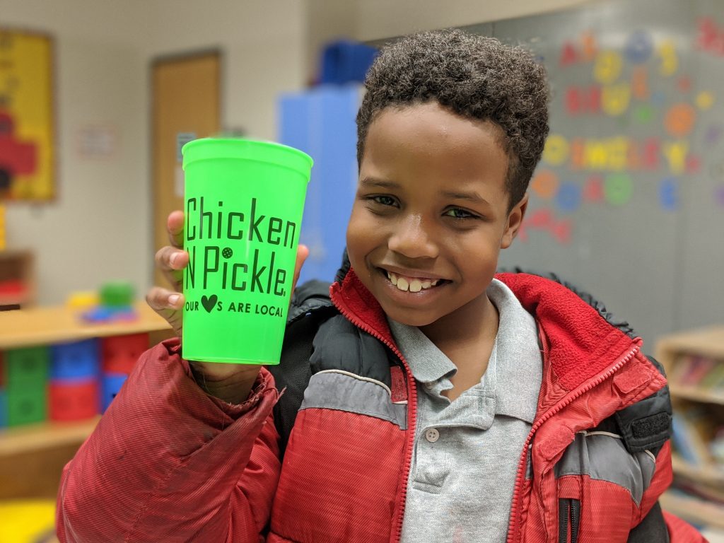 Smiling boy in a red and gray jacket holds up a bright green cup with the words Chicken N Pickle printed on it, standing in a colorful classroom setting.