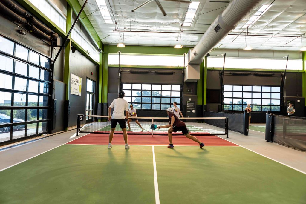 Four people are playing doubles pickleball on an indoor court with green and red flooring. Two players are at the net while two opponents get ready to return the ball. The facility has large windows and bright lighting.