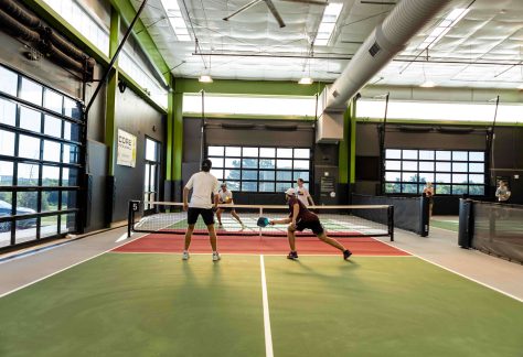 Four people are playing doubles pickleball on an indoor court with green and red flooring. Two players are at the net while two opponents get ready to return the ball. The facility has large windows and bright lighting.