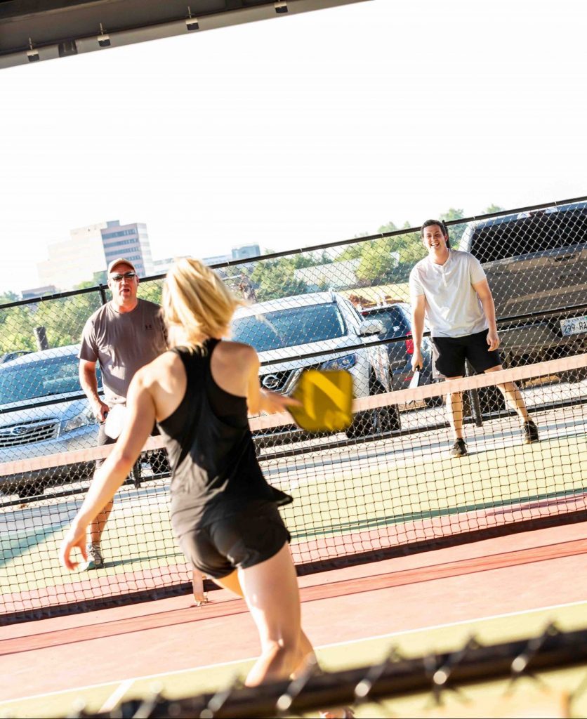 Three people are playing pickleball outdoors on a sunny day. A woman with a yellow paddle faces two men across the net, with cars and buildings visible in the background.