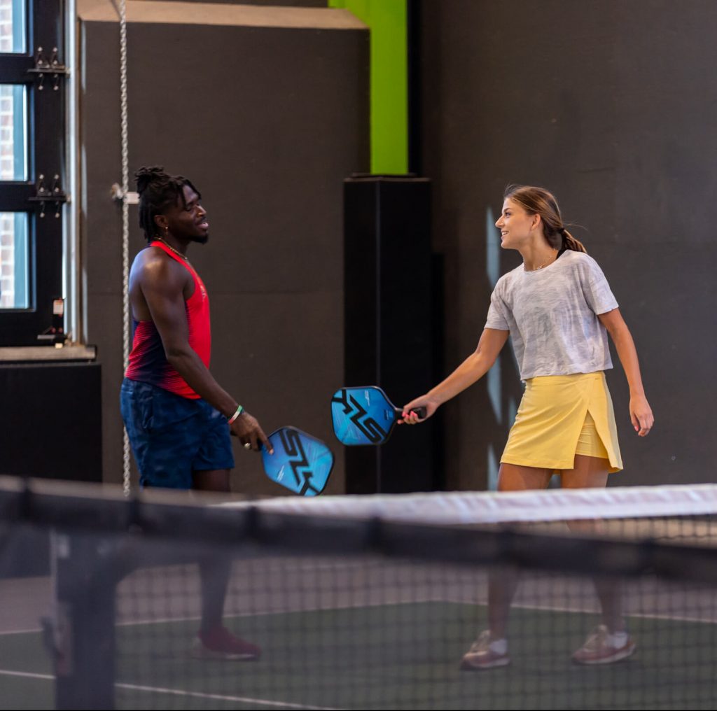 Two people smiling and playing pickleball indoors. The man wears a red tank top and blue shorts, and the woman wears a gray shirt and yellow skirt. Both hold paddles and appear to be enjoying the game.