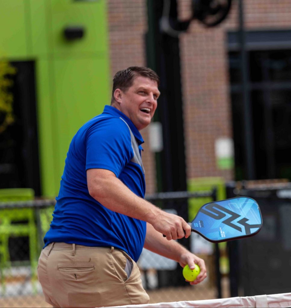 A man in a blue polo shirt and khaki pants is playing pickleball outdoors. He is holding a paddle and preparing to hit a yellow ball, with a fence and buildings in the background.