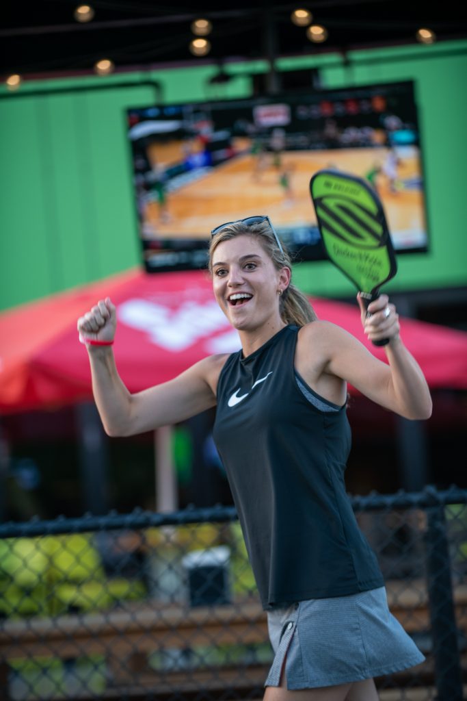 A smiling woman in athletic wear holds a pickleball paddle and raises her arms in celebration. A blurry TV showing a sports game and red umbrellas are visible in the background.