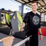 Two people holding Chicken N Pickle paddles tap them together over a pickleball net at an indoor court. One person is in focus, smiling, while the other is in the foreground wearing a smartwatch.