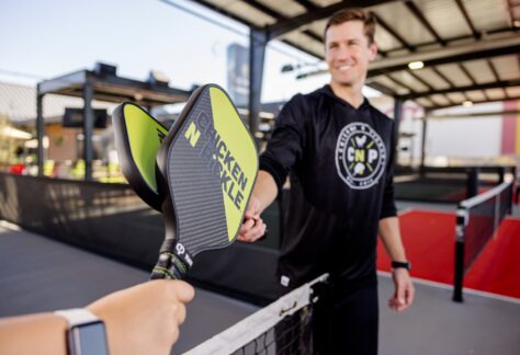 Two people holding Chicken N Pickle paddles tap them together over a pickleball net at an indoor court. One person is in focus, smiling, while the other is in the foreground wearing a smartwatch.