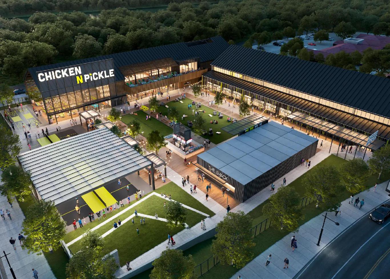 Aerial view of Chicken N Pickle, a modern recreational complex with outdoor seating, pickleball courts, a central green lawn with people, and surrounding buildings, all surrounded by trees and parking areas.
