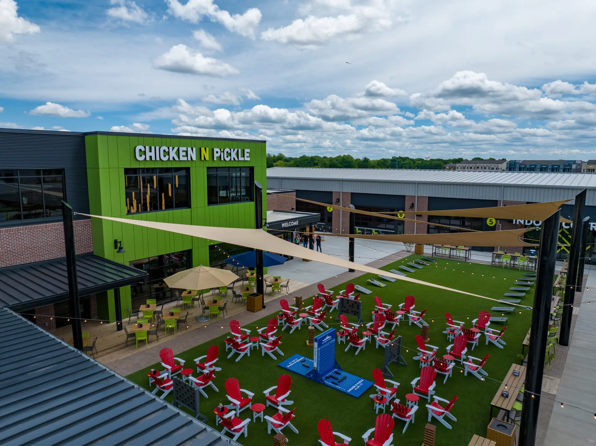 An outdoor area at Chicken N Pickle with green turf, red Adirondack chairs, tables, umbrellas, and yard games, surrounded by modern buildings under a partly cloudy sky.