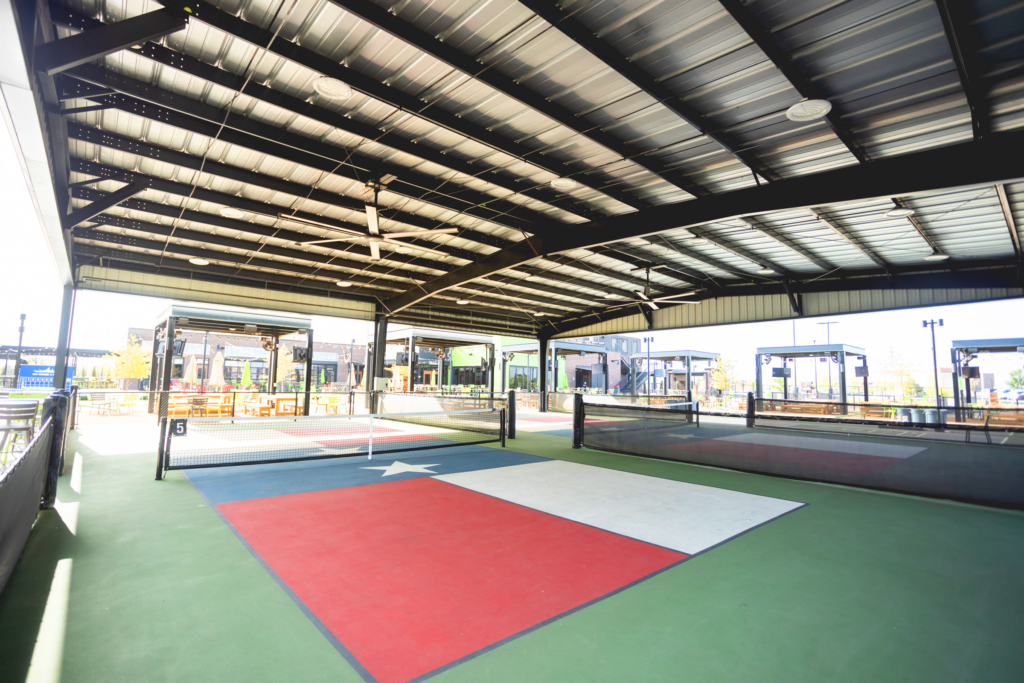 A covered outdoor sports court features a large Texas flag design on the floor, with netted areas and open sides allowing natural light to enter. Buildings and seating areas are visible in the background.