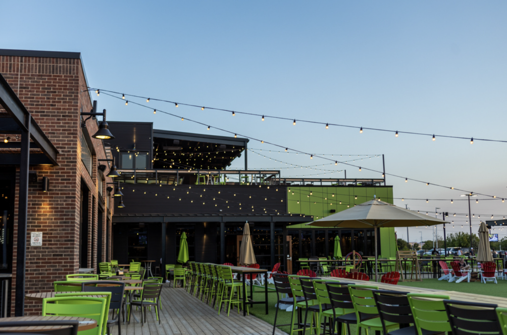 Outdoor patio with green chairs and tables, string lights overhead, and a modern building with brick and dark siding. There are large umbrellas, and the scene is set at dusk with a clear sky.