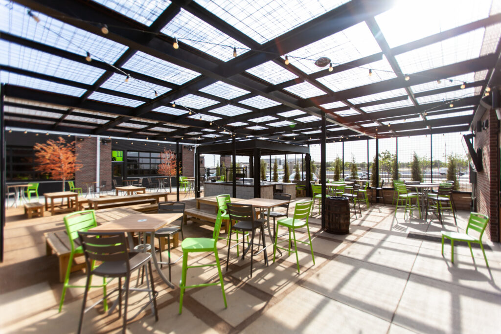 Modern outdoor patio with green chairs and wooden tables under a metal and glass pergola, string lights overhead, and trees visible in the background on a sunny day.