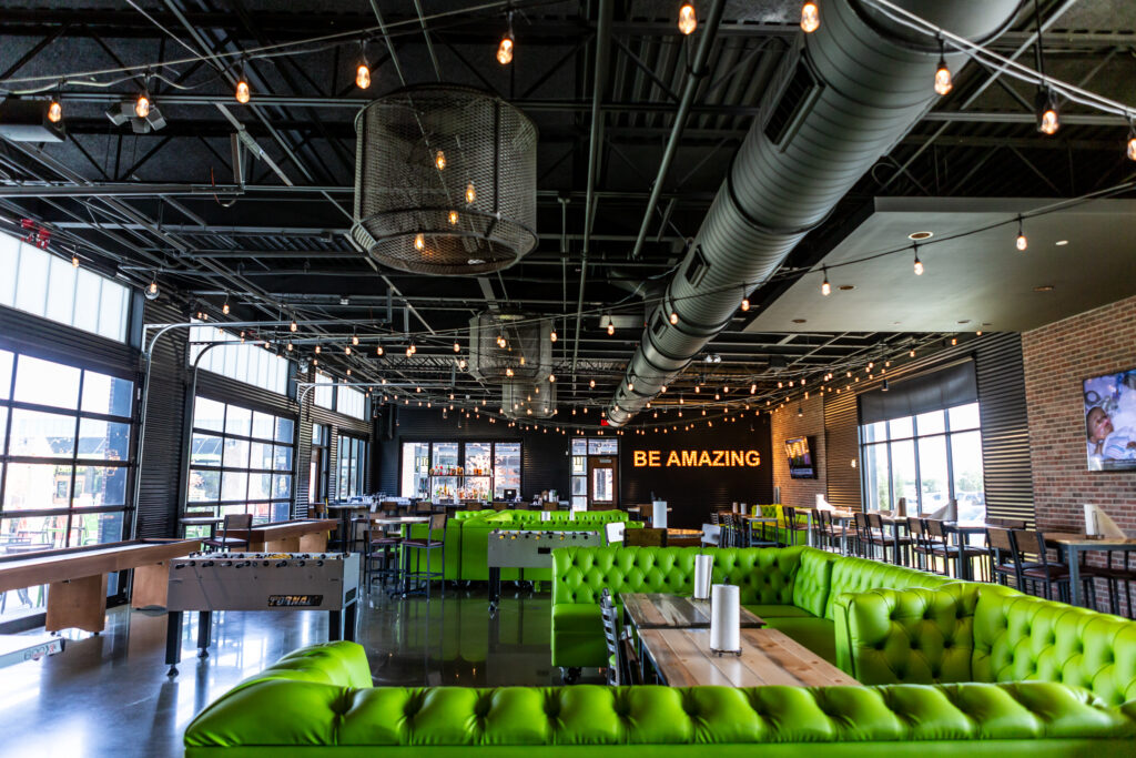 A modern restaurant interior with bright green booth seating, wooden tables, string lights, large windows, and a BE AMAZING sign illuminated on the back wall. The space has an industrial-style ceiling and a polished floor.